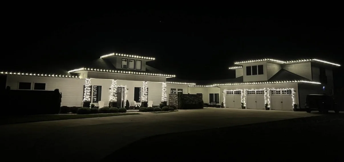 White two-story home at night with Christmas lights along every roofline, wrapped porch columns and outlined garage doors