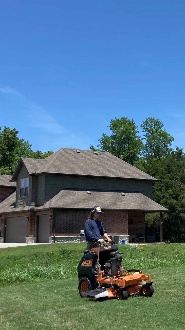 Crew member mowing a lawn on an orange stand-on mower in front of a two-story home