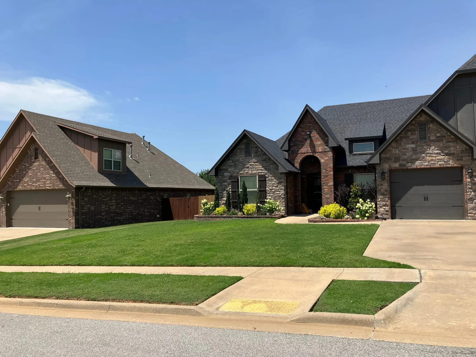 Two brick and stone homes with finished front lawns, trimmed beds and flowering shrubs along the drive