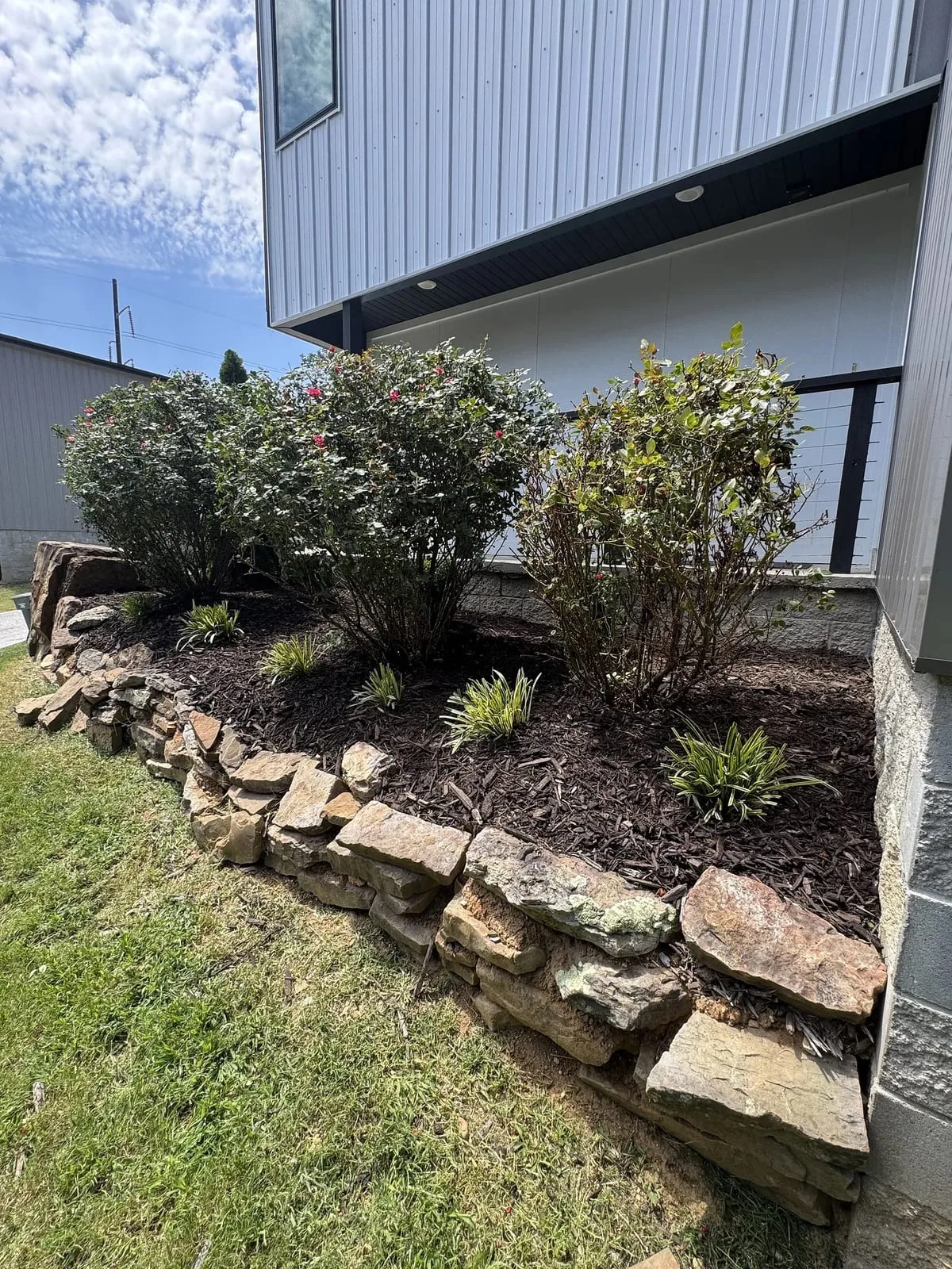 Stacked natural-stone border holding a mulched bed of shrubs beside a modern grey home